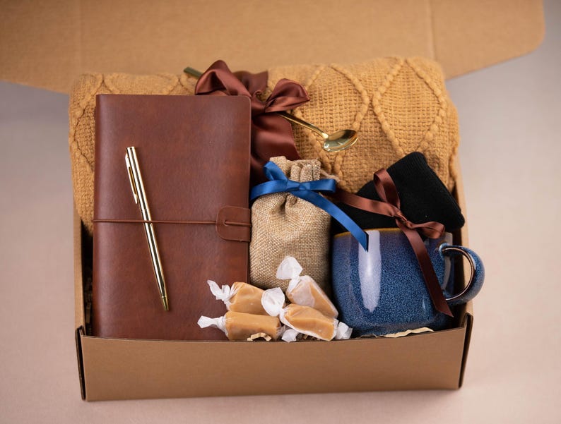 Peut inclure: Une bo&icirc;te en carton marron remplie de divers cadeaux, dont un journal en cuir marron avec un stylo dor&eacute;, une tasse en c&eacute;ramique bleue, un petit sac en toile de jute avec un ruban dor&eacute;, des bonbons au caramel et une couverture en tricot marron.