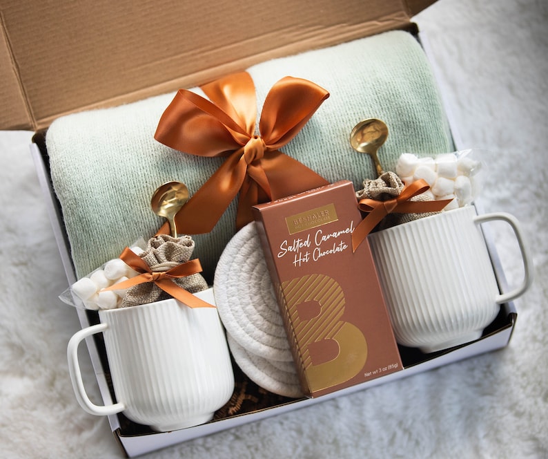 Puede incluir: Una caja de regalo con una manta verde claro, dos tazas blancas y una caja de chocolate caliente con caramelo salado. Las tazas tienen l&iacute;neas verticales y est&aacute;n llenas de malvaviscos. Cucharas doradas y cinta marr&oacute;n.