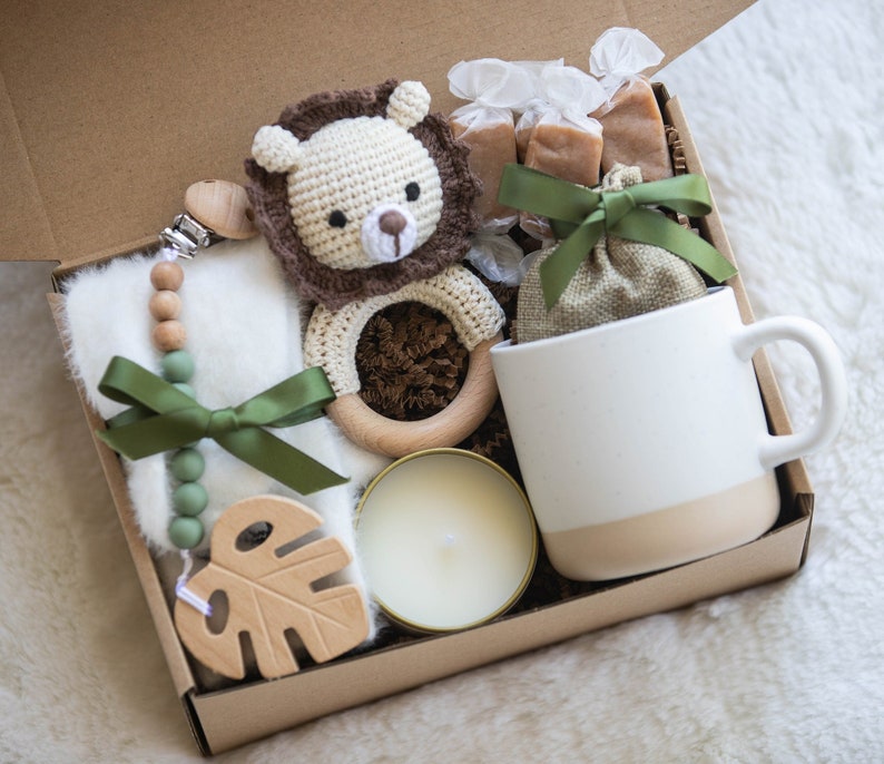May include: A gift box filled with baby items, including a crocheted lion rattle, a wooden teether, a muslin blanket, a candle, and a mug. The box is decorated with green ribbon and a small burlap bag.
