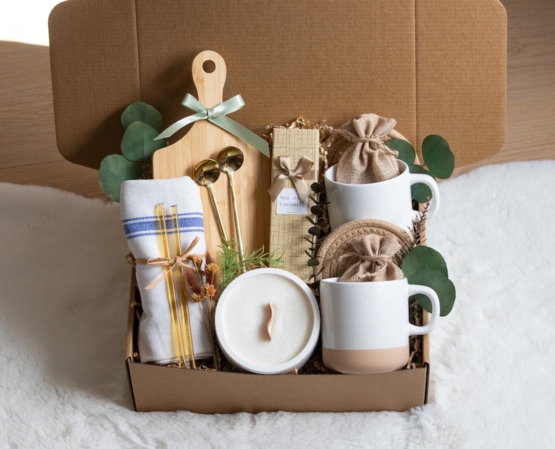 May include: A gift box filled with a wooden cutting board, two white mugs with tan bottoms, a candle, two gold spoons, a linen towel, and other items. The box is brown and contains decorative greenery and small burlap bags.