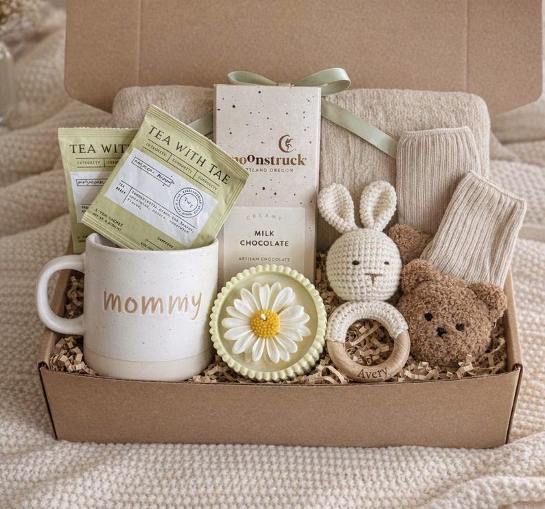 May include: A gift box filled with a variety of items. Includes a mug with "mommy" printed on it, tea bags, chocolate, a crochet bunny, a wooden teether, a teddy bear, a daisy-shaped soap, and socks. The box is tied with a green ribbon.