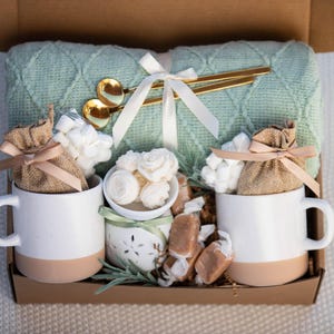 May include: A gift box containing a sage green cable knit throw blanket, two white and tan ceramic mugs, two burlap pouches tied with tan ribbons, a small white ceramic candle holder with floral wax melts, and gold-toned spoons.