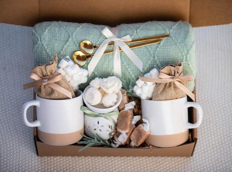May include: A gift box containing a sage green cable knit throw blanket, two white and tan ceramic mugs, two burlap pouches tied with tan ribbons, a small white ceramic candle holder with floral wax melts, and gold-toned spoons.