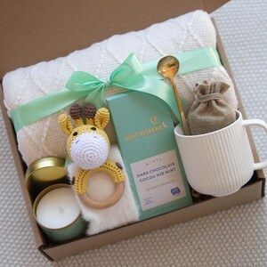May include: A gift box filled with a white knitted blanket, a yellow crocheted giraffe toy, a candle, a mug, a bag of cocoa mix, and a gold spoon. The box is tied with a green ribbon.