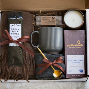 May include: A gift box filled with a brown scarf, a mug, a bag of coffee, a bar of dark chocolate, a small bag of sweets, and a candle. The box is lined with brown paper and the items are arranged neatly.
