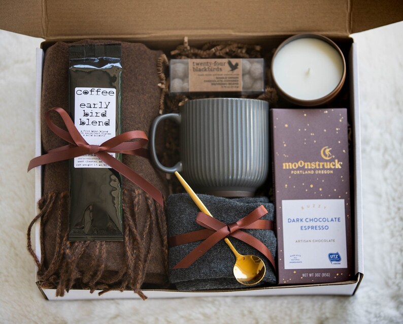 May include: A gift box filled with a brown scarf, a mug, a bag of coffee, a bar of dark chocolate, a small bag of sweets, and a candle. The box is lined with brown paper and the items are arranged neatly.