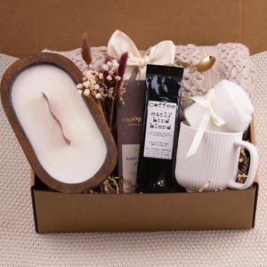 May include: A gift box filled with a wooden candle, coffee, a white mug, and a cozy blanket. The candle is in a wooden bowl. The coffee bag has the text "early bird blend". The mug has vertical lines. A gold spoon and dried flowers are included.