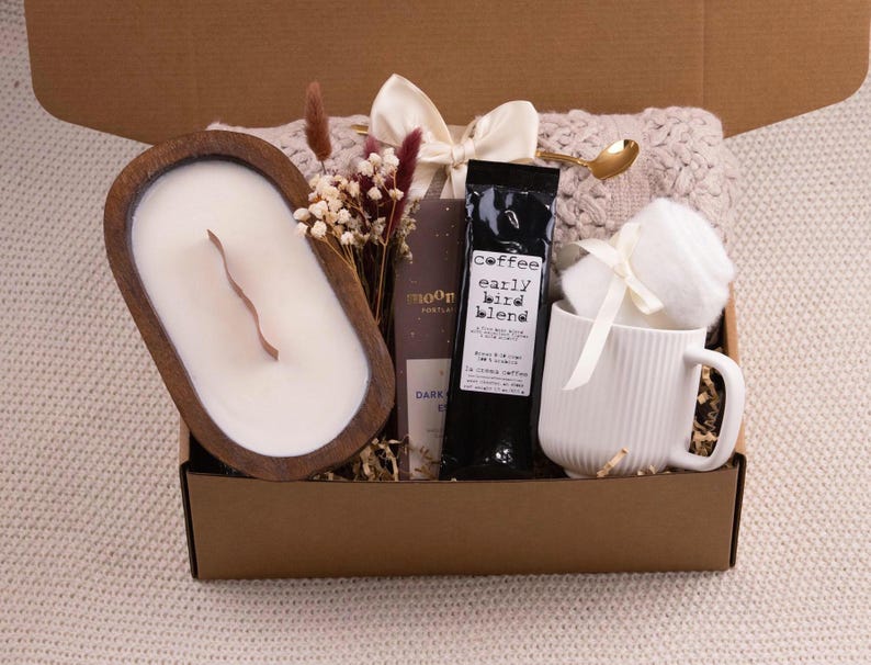 May include: A gift box filled with a wooden candle, coffee, a white mug, and a cozy blanket. The candle is in a wooden bowl. The coffee bag has the text "early bird blend". The mug has vertical lines. A gold spoon and dried flowers are included.