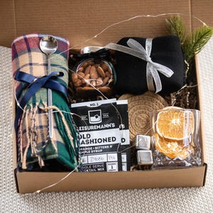 May include: A gift box filled with items for a cosy night in, including a plaid scarf, a jar of almonds, a black sock, a cocktail mix packet, a coaster, and a glass with a dried orange slice.