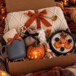 May include: A gift box containing a cream-colored knit blanket with an orange ribbon, a gray ribbed mug with a bow, a small burlap pouch, a bag of marshmallows, a ceramic pumpkin lantern, and a candle decorated with pumpkin-shaped wax.