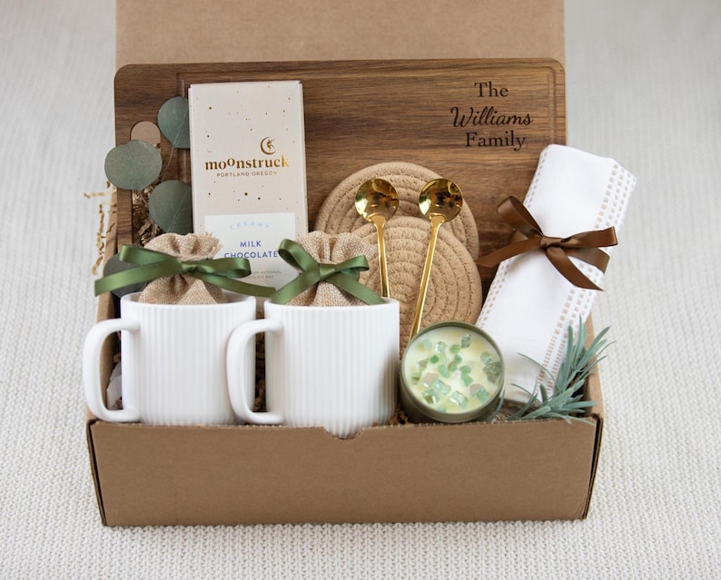 May include: A gift box containing two white ribbed ceramic mugs with burlap pouches tied with green ribbons, a Moonstruck milk chocolate bar, a wooden cutting board engraved with The Williams Family, two gold spoons, coasters, and a candle.