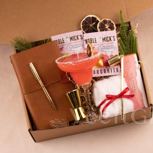 May include: A gift box filled with cocktail-themed items. It includes a margarita cocktail, a leather-bound journal with a gold pen, cocktail mix packets, a jigger, and a small white cloth with a red ribbon.