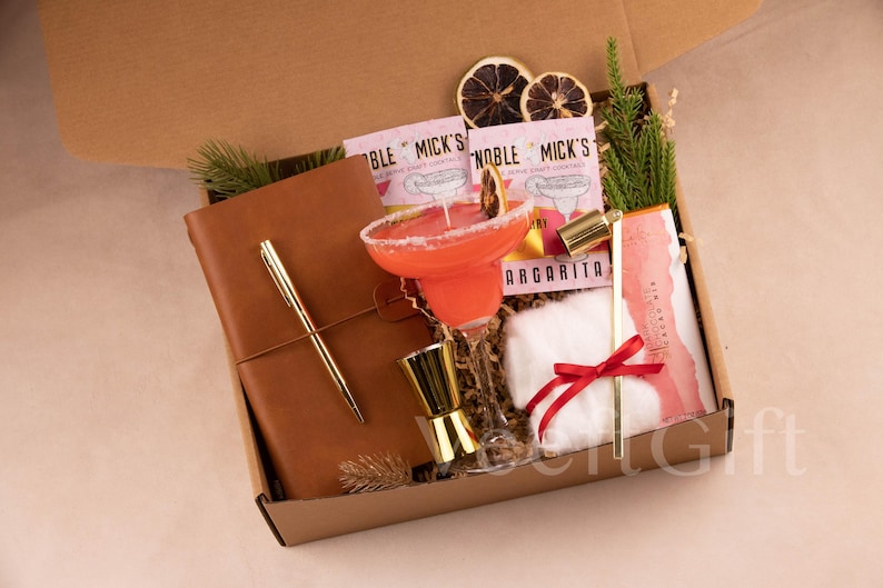 May include: A gift box filled with cocktail-themed items. It includes a margarita cocktail, a leather-bound journal with a gold pen, cocktail mix packets, a jigger, and a small white cloth with a red ribbon.