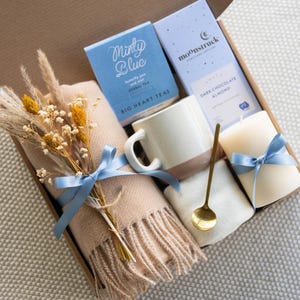 K&ouml;nnte beinhalten: Eine Geschenkbox mit einem beigen Schal, einer Tasse, Tee, Schokolade und einer Kerze, alle mit blauen B&auml;ndern gebunden. Die Box enth&auml;lt einen Strau&szlig; Trockenblumen und einen goldenen L&ouml;ffel. Auf der Teebox steht "Minty Blue".