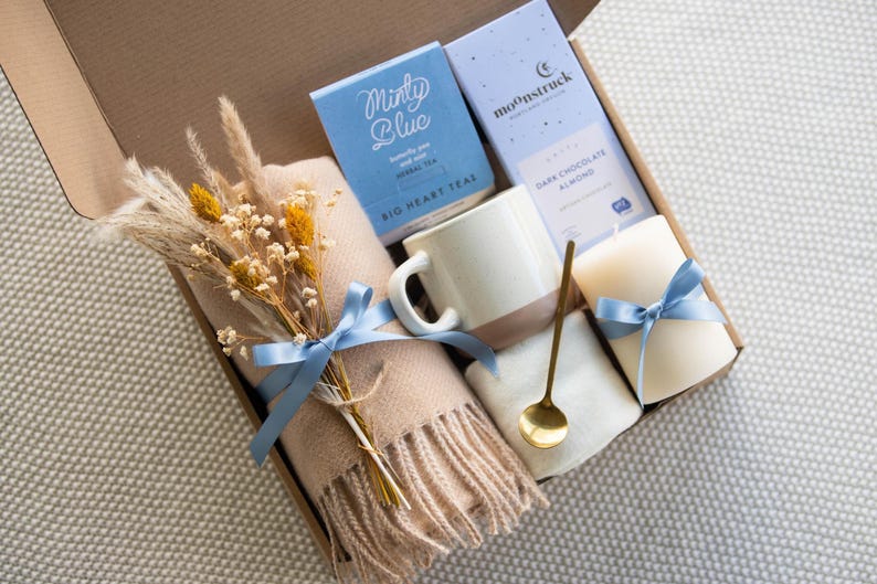K&ouml;nnte beinhalten: Eine Geschenkbox mit einem beigen Schal, einer Tasse, Tee, Schokolade und einer Kerze, alle mit blauen B&auml;ndern gebunden. Die Box enth&auml;lt einen Strau&szlig; Trockenblumen und einen goldenen L&ouml;ffel. Auf der Teebox steht "Minty Blue".