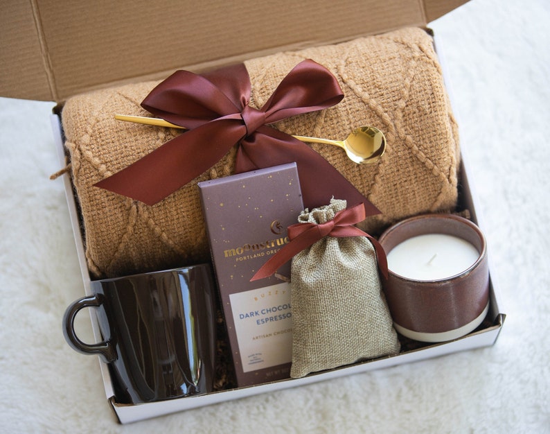 Peut inclure: Une bo&icirc;te-cadeau remplie d'articles confortables pour prendre soin de soi, comprenant une couverture en tricot marron, une tasse en c&eacute;ramique marron, une barre de chocolat noir expresso, un petit sac en toile de jute avec un ruban marron et une bougie en c&eacute;ramique marron.
