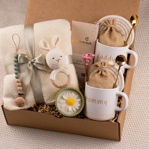 May include: A gift box filled with baby items and treats. Includes a cream-colored blanket, a crochet bunny, a pacifier clip, a wooden teether, two mugs that say "daddy" and "mommy", gold spoons, and chocolate.