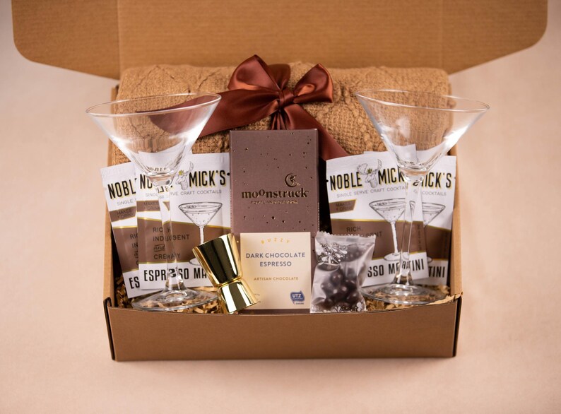 May include: A gift box containing two clear martini glasses, a gold-toned jigger, a brown knit textile, a brown satin ribbon, four Noble Mick's Espresso Martini drink mix packets, a Moonstruck dark chocolate espresso bar, and a small bag of chocolate.