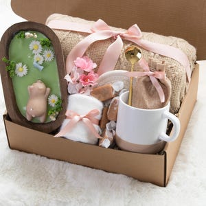 May include: A gift box filled with self-care items. Includes a wooden framed soap with a pig design, a beige blanket tied with a pink ribbon, a white and beige mug, a gold spoon, and other items.