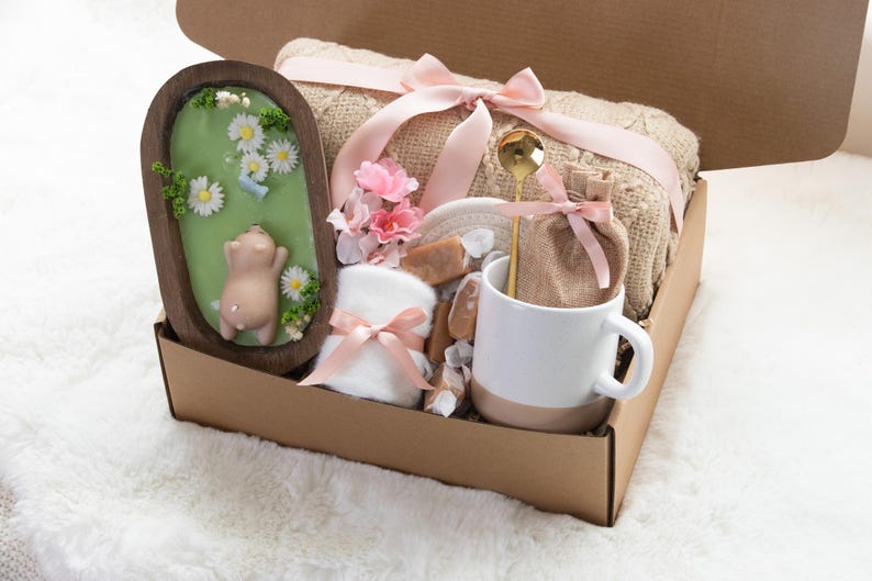 May include: A gift box filled with self-care items. Includes a wooden framed soap with a pig design, a beige blanket tied with a pink ribbon, a white and beige mug, a gold spoon, and other items.