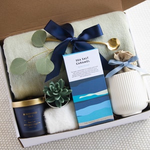Peut inclure: Une bo&icirc;te-cadeau remplie d'une vari&eacute;t&eacute; d'articles, dont une bougie bleue et dor&eacute;e, une petite plante succulente, une tasse blanche, une barre de chocolat avec le texte "Sea Salt Caramel", une couverture en laine bleue et blanche et une cuill&egrave;re dor&eacute;e.