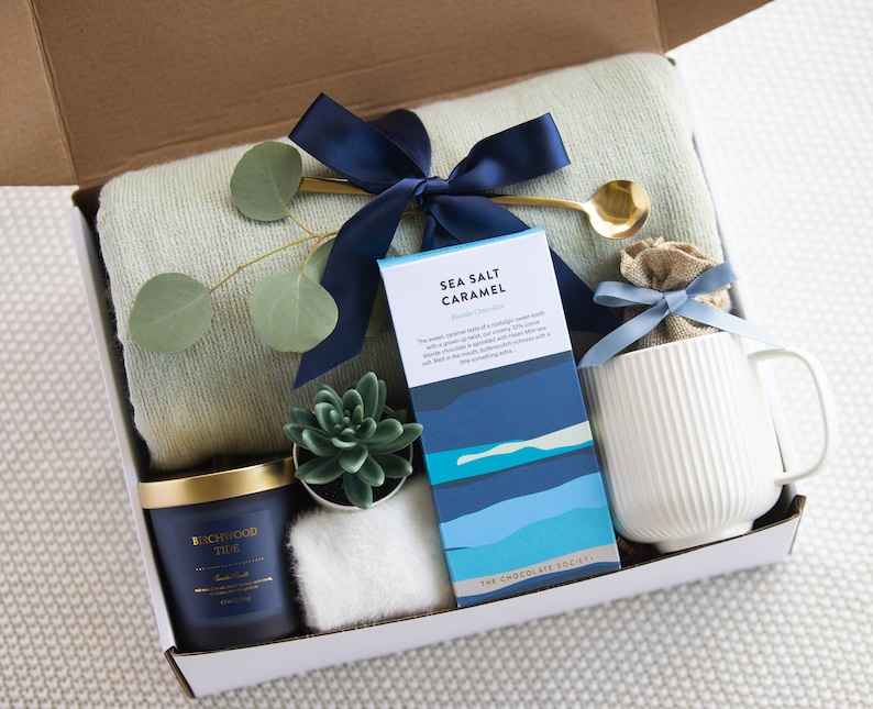 Peut inclure: Une bo&icirc;te-cadeau remplie d'une vari&eacute;t&eacute; d'articles, dont une bougie bleue et dor&eacute;e, une petite plante succulente, une tasse blanche, une barre de chocolat avec le texte "Sea Salt Caramel", une couverture en laine bleue et blanche et une cuill&egrave;re dor&eacute;e.