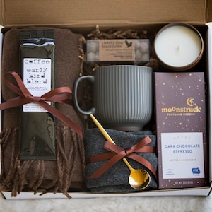 K&ouml;nnte beinhalten: Eine Geschenkbox mit einem braunen Schal, Kaffee, einer grauen Tasse, einer Kerze, Schokolade und einem goldenen L&ouml;ffel. Die Kaffeepackung tr&auml;gt die Aufschrift "early bird blend". Die Schokoladentafel ist von Moonstruck Portland Oregon.