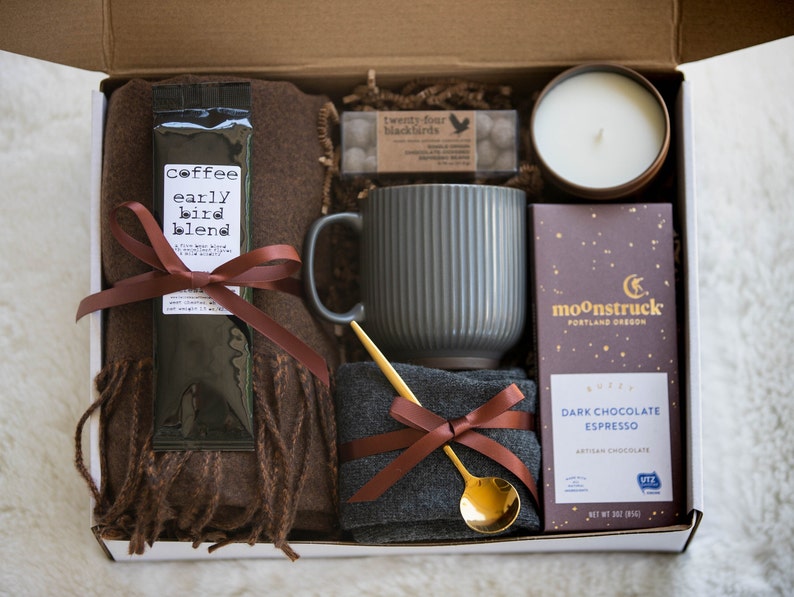 K&ouml;nnte beinhalten: Eine Geschenkbox mit einem braunen Schal, Kaffee, einer grauen Tasse, einer Kerze, Schokolade und einem goldenen L&ouml;ffel. Die Kaffeepackung tr&auml;gt die Aufschrift "early bird blend". Die Schokoladentafel ist von Moonstruck Portland Oregon.