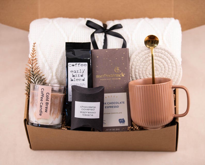 May include: A gift box filled with a white blanket, coffee, chocolate, a candle, and a mug. The mug has a gold spoon inside. The box also contains a small decorative pine branch and a woven coaster.