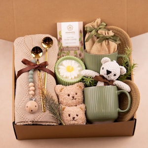 May include: A gift box filled with a beige knitted blanket, gold spoons, a pacifier, coffee, a daisy candle, two teddy bear soaps, two green mugs, a knitted teddy bear, and a woven hat. The box is brown.