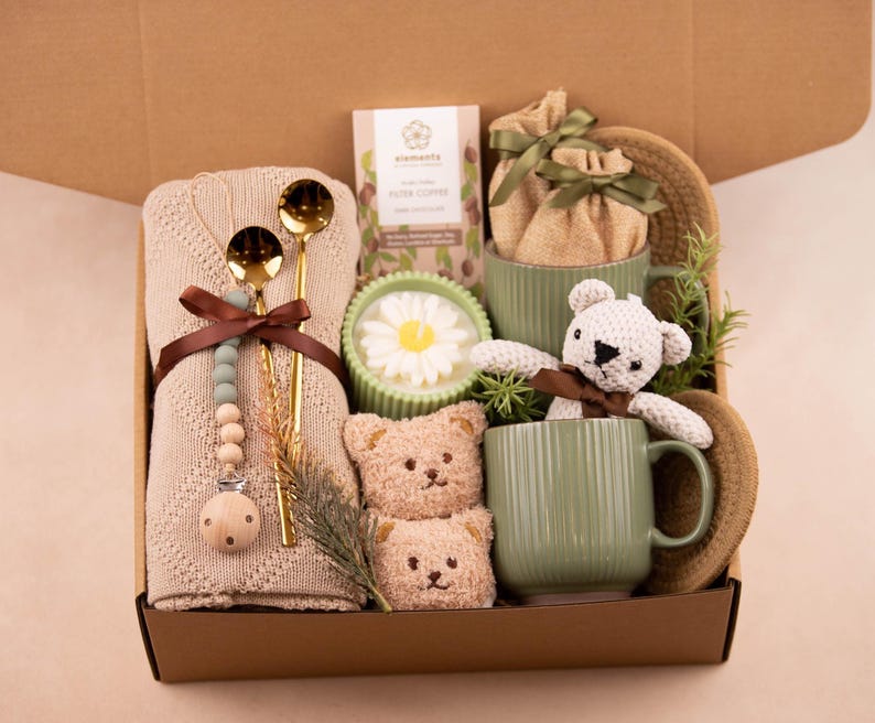 May include: A gift box filled with a beige knitted blanket, gold spoons, a pacifier, coffee, a daisy candle, two teddy bear soaps, two green mugs, a knitted teddy bear, and a woven hat. The box is brown.