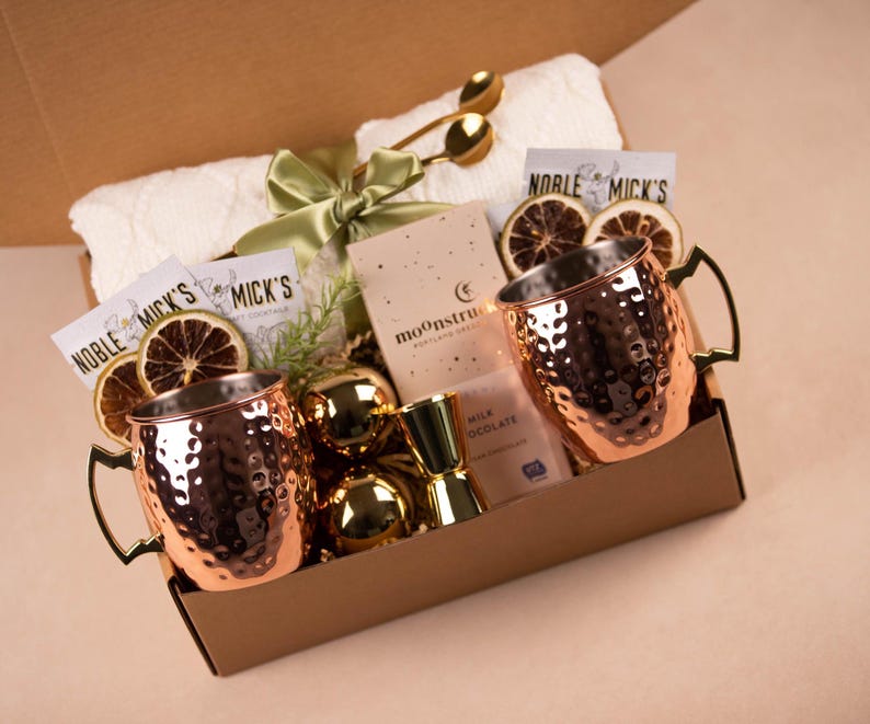 May include: A brown gift box filled with a variety of items, including two copper mugs, a small bottle of milk chocolate, a white towel, dried citrus slices, and gold-colored ornaments. The box is labeled "Noble Mick's" and "Moonstruck".