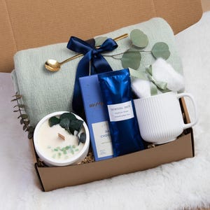 May include: A gift box filled with self-care items. It includes a light green blanket, a white mug, a candle, dark chocolate, and a gold spoon. The items are arranged in a brown box, tied with a navy blue ribbon.