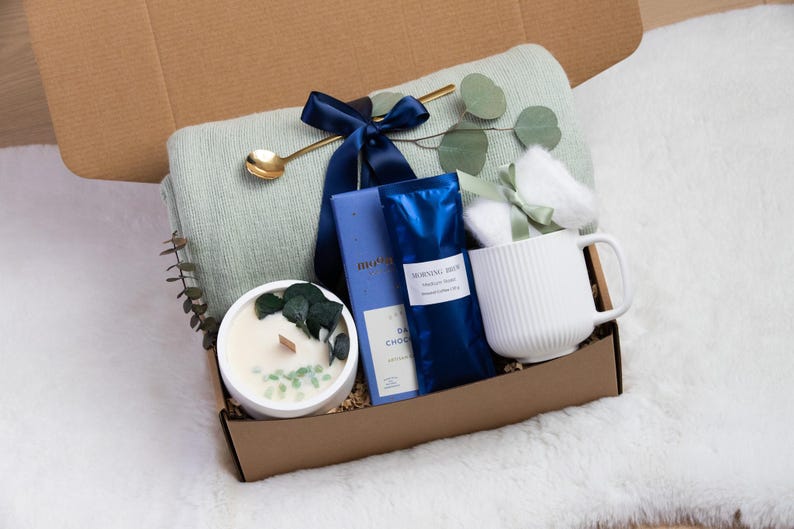 May include: A gift box filled with self-care items. It includes a light green blanket, a white mug, a candle, dark chocolate, and a gold spoon. The items are arranged in a brown box, tied with a navy blue ribbon.