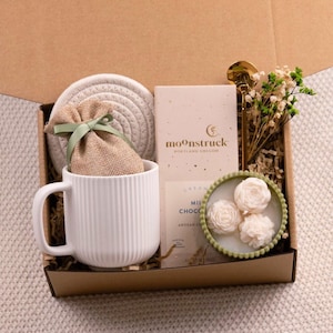 K&ouml;nnte beinhalten: Eine Geschenkbox mit einer wei&szlig;en Tasse, einem Untersetzer, einem kleinen Leinensack mit gr&uuml;ner Schleife, einer Tafel Schokolade und einer dekorativen Schale mit drei wei&szlig;en bl&uuml;tenf&ouml;rmigen Seifen. Ein goldener L&ouml;ffel und Trockenblumen sind ebenfalls enthalten.