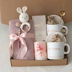 May include: A gift box filled with baby items and gifts for parents. Includes a pink knitted blanket, a crochet bunny, a pacifier, two mugs that say "daddy" and "mommy", a candle, chocolate, and gold spoons.