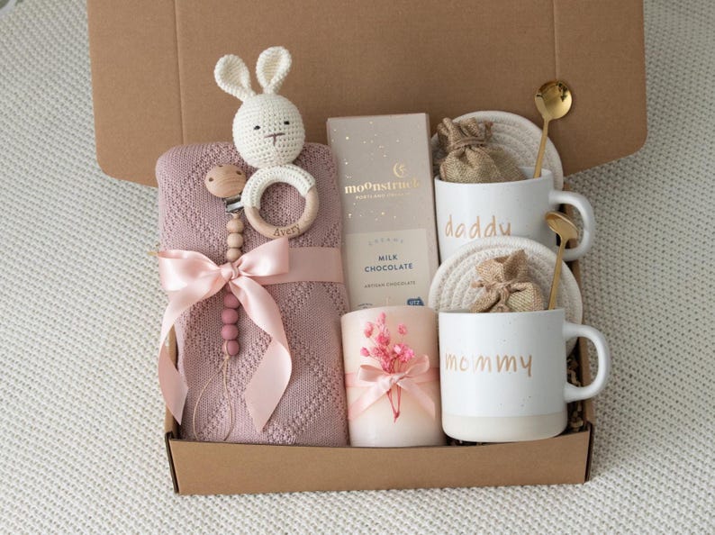 May include: A gift box filled with baby items and gifts for parents. Includes a pink knitted blanket, a crochet bunny, a pacifier, two mugs that say "daddy" and "mommy", a candle, chocolate, and gold spoons.