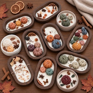 May include: A collection of oval and round candles in wooden and ceramic bowls, featuring white wax topped with decorative wax pumpkins, dried orange slices, star anise, and cinnamon sticks, arranged on a wooden surface with autumn leaves.