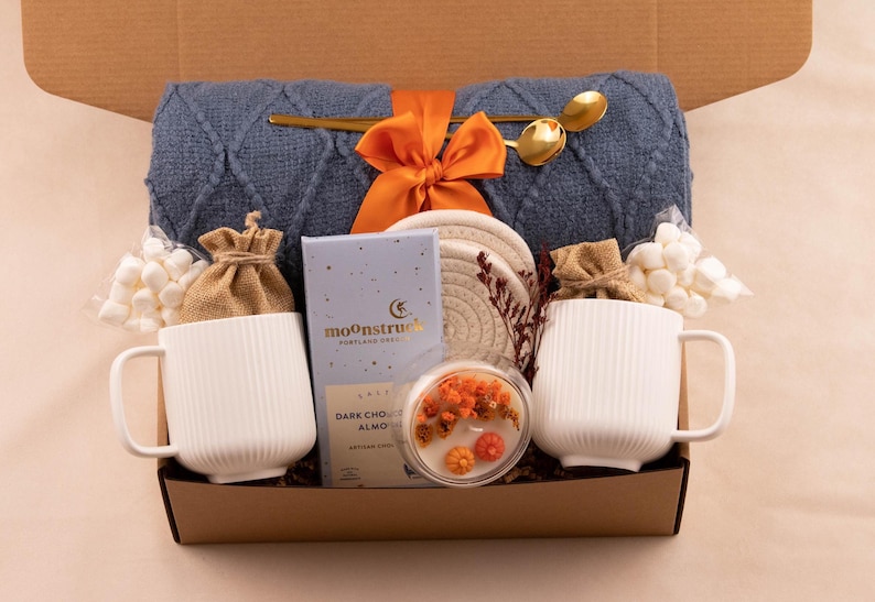 Puede incluir: Una caja de regalo llena de art&iacute;culos acogedores. Incluye una manta de punto azul, dos tazas blancas, una vela, chocolate, malvaviscos y cucharas doradas. Un lazo naranja adorna la manta. La caja es marr&oacute;n.
