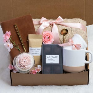 May include: A gift box filled with a brown leather journal, gold pen, candle, coffee, dark chocolate caramels, a mug, and a blanket tied with pink ribbons. Pink flowers and a gold spoon complete the arrangement.