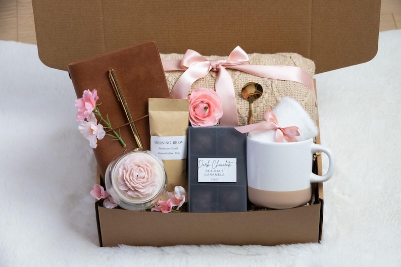 May include: A gift box filled with a brown leather journal, gold pen, candle, coffee, dark chocolate caramels, a mug, and a blanket tied with pink ribbons. Pink flowers and a gold spoon complete the arrangement.