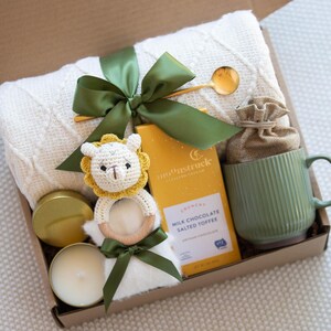 Op de afbeelding: Een cadeaudoos gevuld met artikelen voor een pasgeboren baby, waaronder een wit gebreid dekentje, een gele en groene gehaakte dierenrammelaar, een kaars, een mok en een doos met melkchocolade met gezouten toffee.