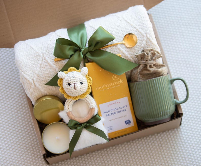 Op de afbeelding: Een cadeaudoos gevuld met artikelen voor een pasgeboren baby, waaronder een wit gebreid dekentje, een gele en groene gehaakte dierenrammelaar, een kaars, een mok en een doos met melkchocolade met gezouten toffee.