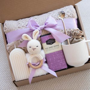 May include: A gift box filled with items for a new baby, including a knitted blanket, a crocheted bunny rattle, a candle, a bar of chocolate, a mug, and a spoon.
