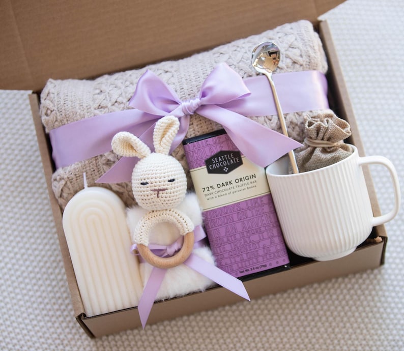 May include: A gift box filled with items for a new baby, including a knitted blanket, a crocheted bunny rattle, a candle, a bar of chocolate, a mug, and a spoon.