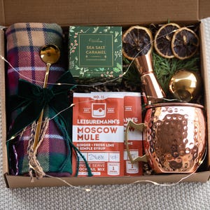 May include: A gift box filled with items for a cosy night in, including a plaid throw blanket, a copper mug, a cocktail mix packet, a bottle of sea salt caramel, and a jigger.