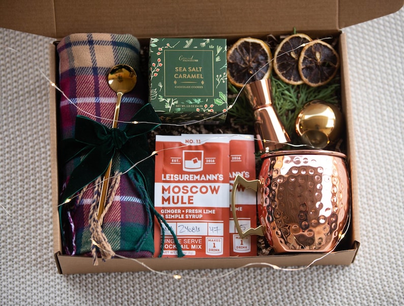 May include: A gift box filled with items for a cosy night in, including a plaid throw blanket, a copper mug, a cocktail mix packet, a bottle of sea salt caramel, and a jigger.