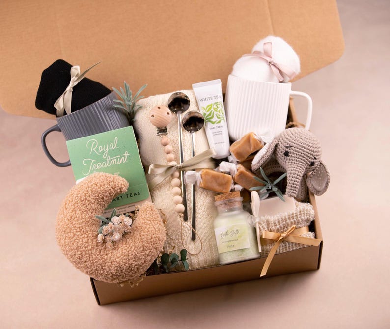 May include: A gift box filled with self-care items. Includes a gray mug, a black sleep mask, a green tea box with the text "Royal Treatment", a white mug, a small crocheted elephant, and other items.