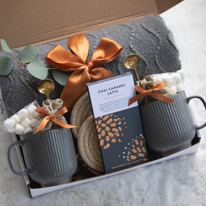 Puede incluir: Una caja de regalo con una manta gris de punto trenzado, dos tazas grises acanaladas, una tableta de chocolate Chai Caramel Latte y peque&ntilde;as bolsas de malvaviscos. La caja est&aacute; atada con una cinta de raso naranja e incluye cucharas doradas y un posavasos tejido.
