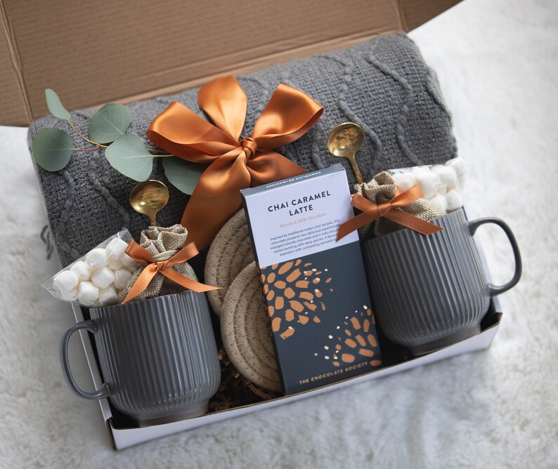 Puede incluir: Una caja de regalo con una manta gris de punto trenzado, dos tazas grises acanaladas, una tableta de chocolate Chai Caramel Latte y peque&ntilde;as bolsas de malvaviscos. La caja est&aacute; atada con una cinta de raso naranja e incluye cucharas doradas y un posavasos tejido.